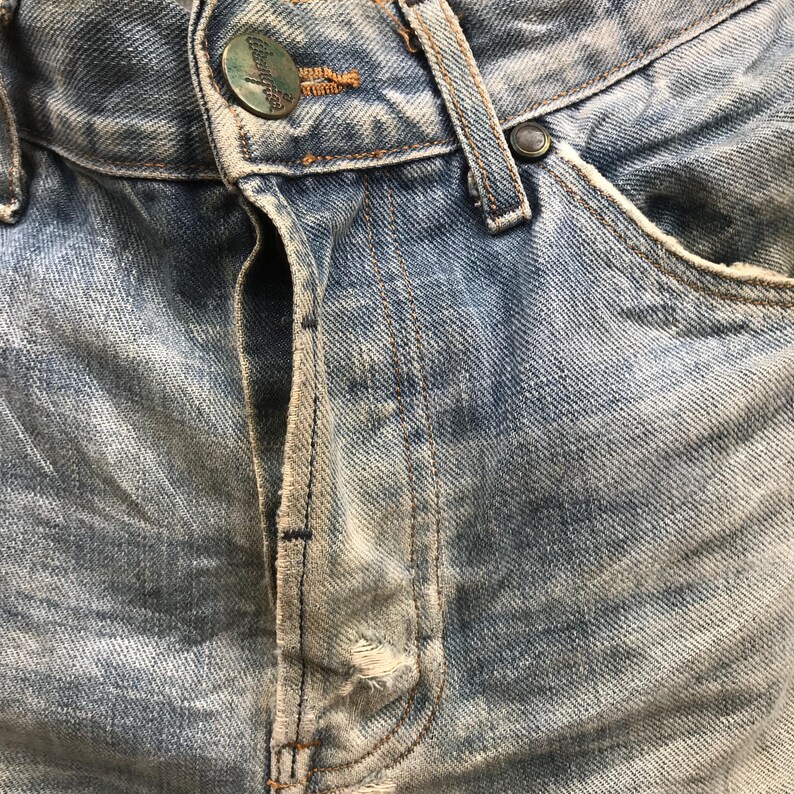 May include: Close-up of light blue denim jeans. The jeans show wear and tear, with visible fading and distressing. A metal button and a small pocket are visible. The jeans have a classic five-pocket design.