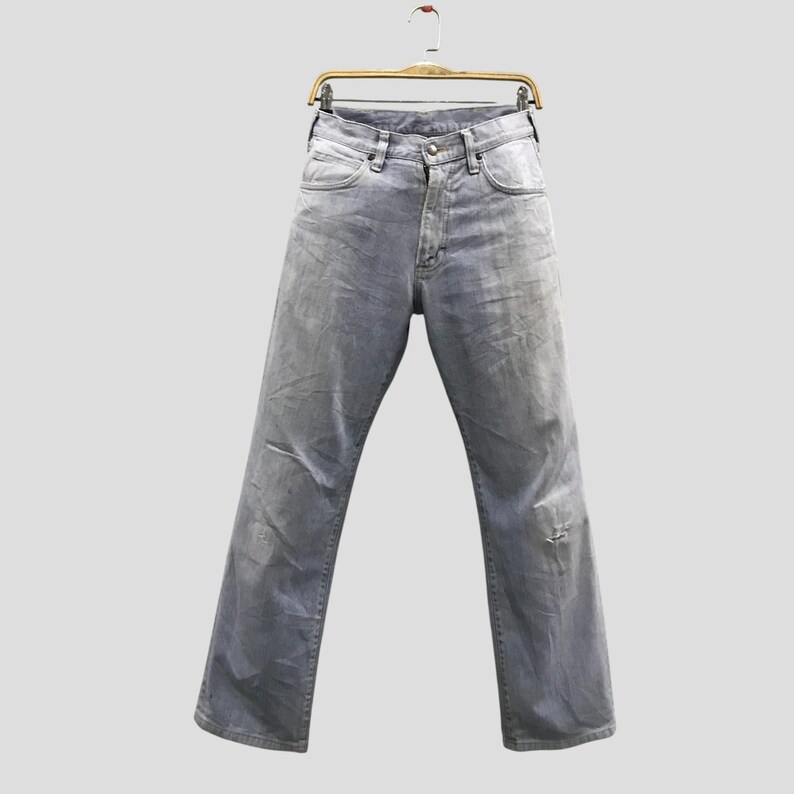 May include: Light grey denim jeans hanging on a wooden hanger. The jeans have a classic five-pocket design, a button and zip closure, and a distressed look with a small tear on the right leg.
