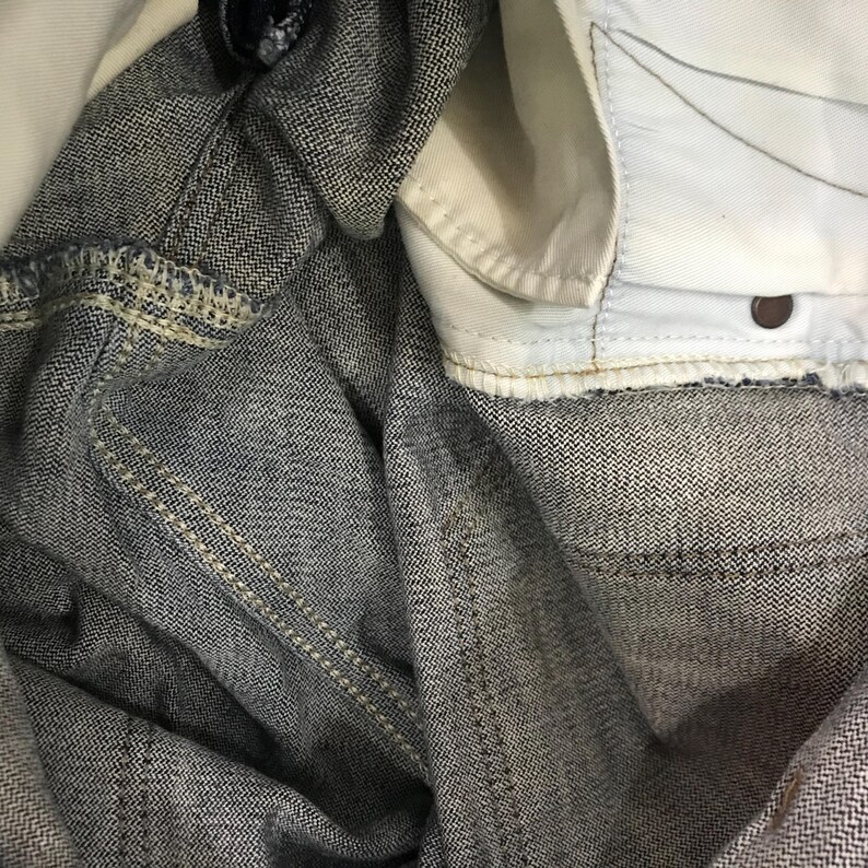 May include: Close-up of denim jeans fabric. The jeans are a medium wash blue with a subtle herringbone weave pattern. The interior lining is white, and the stitching is a contrasting beige colour.