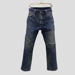 May include: Faded blue denim jeans hanging on a gold-colored hanger. The jeans have a button fly and show signs of wear, with lighter patches on the knees and thighs. The jeans are a classic, straight-leg style.