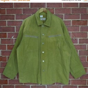 May include: A green suede jacket with a button-up front and two zipped pockets on the chest. The jacket has a relaxed fit and long sleeves.