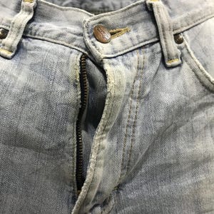 May include: Close-up of light blue denim jeans. The jeans have a metal zip, a button with the brand name "Wrangler", and visible stitching. The denim has a faded, worn appearance.