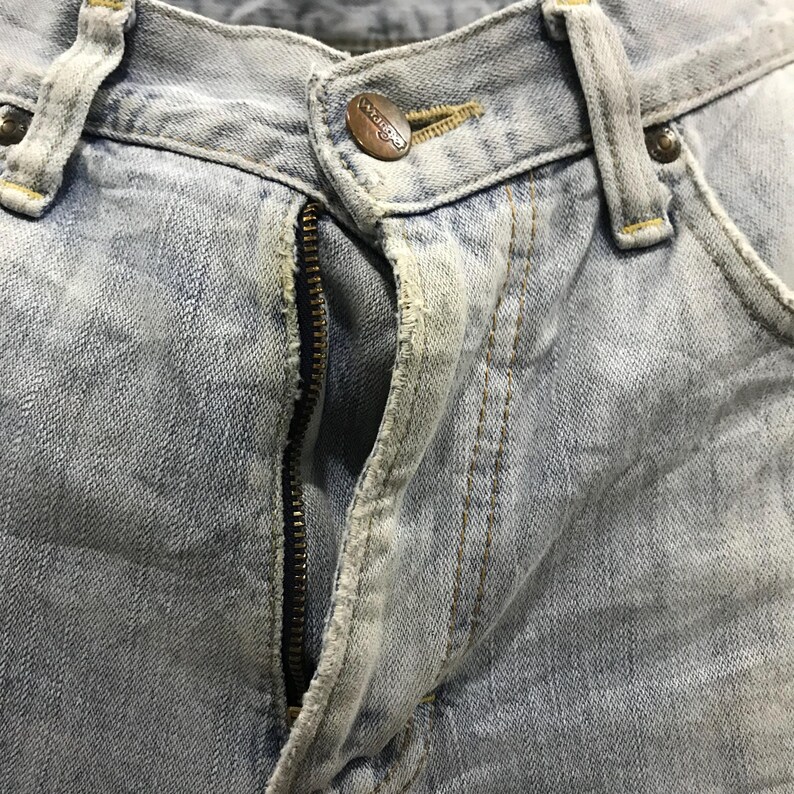 May include: Close-up of light blue denim jeans. The jeans have a metal zip, a button with the brand name "Wrangler", and visible stitching. The denim has a faded, worn appearance.