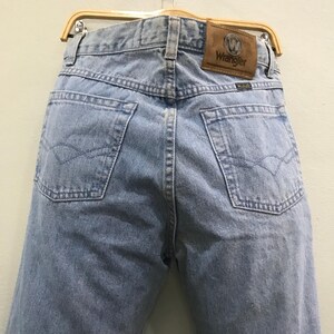 May include: Light blue denim jeans with a classic five-pocket design. The back features two pockets with a decorative stitch and a brown leather patch with the "Wrangler" logo. The jeans are hanging on a wooden hanger.