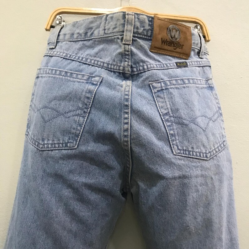 May include: Light blue denim jeans with a classic five-pocket design. The back features two pockets with a decorative stitch and a brown leather patch with the "Wrangler" logo. The jeans are hanging on a wooden hanger.