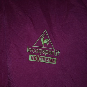 Puede incluir: Una chaqueta color burdeos con un logotipo bordado en verde que dice "le coq sportif" y "NEXTREME" en un rect&aacute;ngulo.