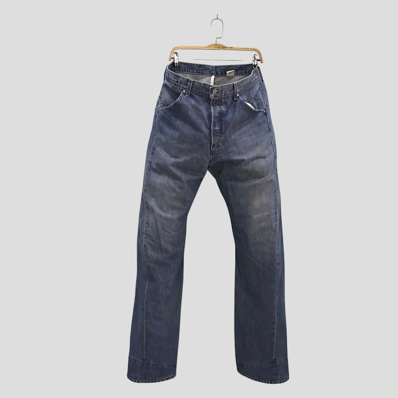 May include: A pair of blue denim jeans featuring a classic five-pocket design, a button fly closure, and a faded wash finish. The jeans are displayed hanging from a wooden hanger, showcasing a straight-leg cut with visible stitching details.