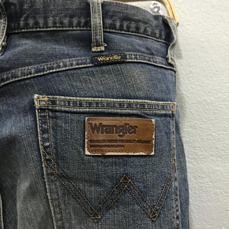 Pu&ograve; includere: Primo piano del retro di jeans blu. I jeans presentano una toppa in pelle marrone con il logo e il testo "Wrangler". La tasca posteriore ha un caratteristico design con cucitura a "W". I jeans sono appesi a una gruccia di legno.