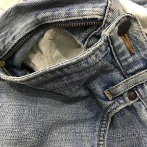 May include: Close-up of light blue denim jeans, showing the zipper, button, and pocket details. The jeans have a worn, faded appearance, with visible stitching and a metal zipper.