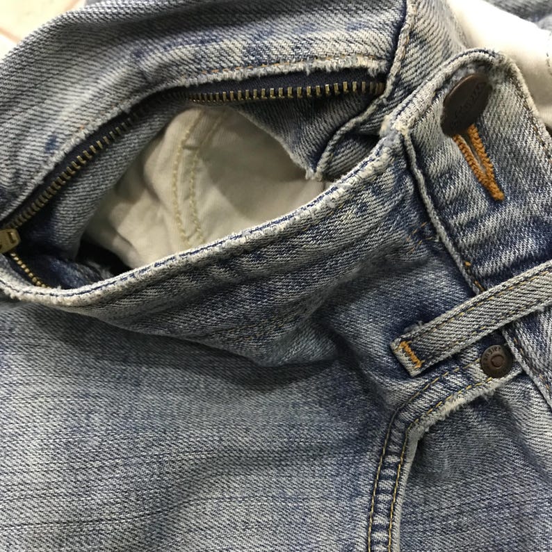 May include: Close-up of light blue denim jeans, showing the zipper, button, and pocket details. The jeans have a worn, faded appearance, with visible stitching and a metal zipper.