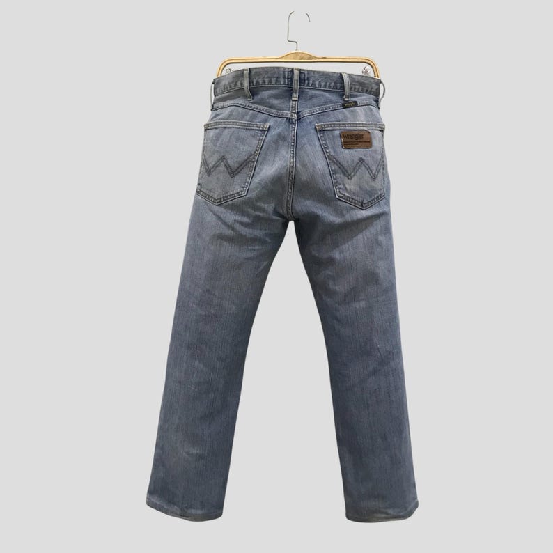 May include: Light blue denim jeans with the Wrangler logo on the back pocket. The jeans are hanging on a wooden hanger. The jeans have a classic five-pocket design and a straight leg.