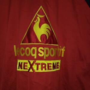 Puede incluir: Chaqueta roja con un logotipo de gallo bordado en amarillo y el texto "le coq sportif" y "NEXTREME".