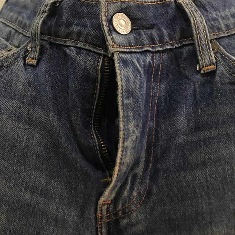 May include: Close-up of blue denim jeans with a partially open zipper. The jeans have a button closure and visible stitching in a contrasting orange-brown thread. The fabric shows signs of wear.