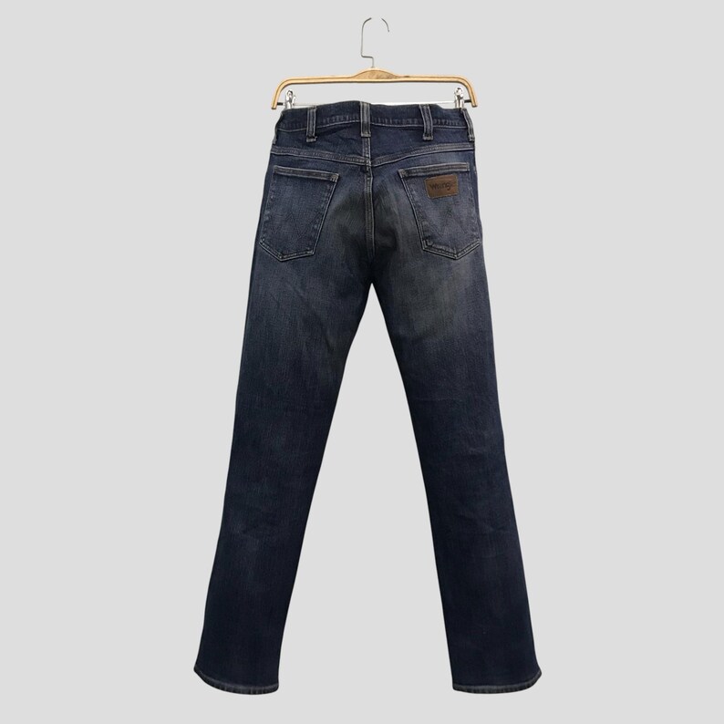 May include: Dark wash denim jeans hanging on a wooden hanger. The jeans feature a classic five-pocket design, a brown leather patch, and a straight leg. The back view of the jeans is shown.