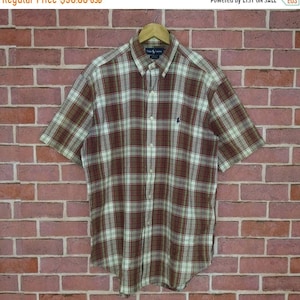 Puede incluir: Una camisa de manga corta con botones de cuadros, con un patrón de cuadros marrones, blancos y verdes. La camisa tiene un pequeño logotipo bordado en el pecho izquierdo.