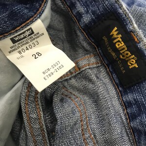 Op de afbeelding: Close-up van een blauwe spijkerbroek met een wit label met de tekst "Wrangler" en "Maat 28". Het label bevat ook de tekst "W04033", "WCM-2537" en "E789-2103". Een zwart label met gouden tekst "Wrangler" en "GREENSBORO, N.C. U.S.A." is ook zichtbaar.