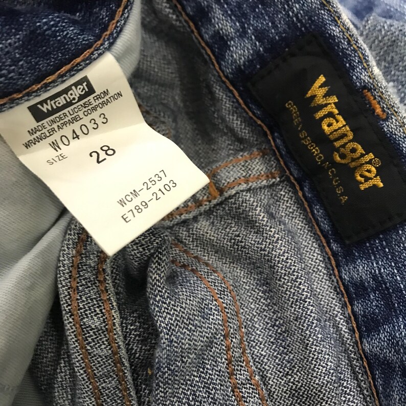 Op de afbeelding: Close-up van een blauwe spijkerbroek met een wit label met de tekst "Wrangler" en "Maat 28". Het label bevat ook de tekst "W04033", "WCM-2537" en "E789-2103". Een zwart label met gouden tekst "Wrangler" en "GREENSBORO, N.C. U.S.A." is ook zichtbaar.