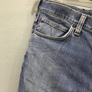 May include: Close-up of light blue denim jeans. The jeans have a classic five-pocket design, with a small pocket on the front and a visible coin pocket. The denim shows signs of wear and fading, with a metal button and rivets.