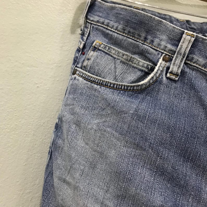 May include: Close-up of light blue denim jeans. The jeans have a classic five-pocket design, with a small pocket on the front and a visible coin pocket. The denim shows signs of wear and fading, with a metal button and rivets.