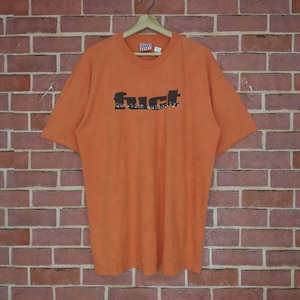 May include: Orange t-shirt with the word "fuct" in a bold, stylized font, and the words "Question authority." below it. The shirt is hanging on a wooden hanger against a brick wall.