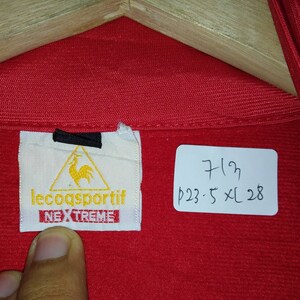 Puede incluir: Chaqueta roja Le Coq Sportif con un logotipo negro y amarillo y el texto "Le Coq Sportif" y "Nextreme". La etiqueta est&aacute; unida a una etiqueta blanca. Una etiqueta blanca con texto negro dice "7/23 p23:5 XL 28".