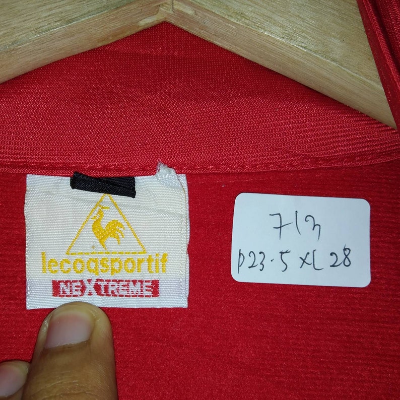 Puede incluir: Chaqueta roja Le Coq Sportif con un logotipo negro y amarillo y el texto "Le Coq Sportif" y "Nextreme". La etiqueta est&aacute; unida a una etiqueta blanca. Una etiqueta blanca con texto negro dice "7/23 p23:5 XL 28".