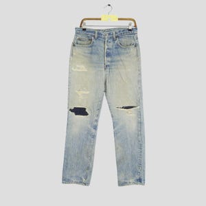 May include: Light blue denim jeans with distressed details, including rips at the knees and thighs. The jeans are hanging on a wooden hanger. The jeans have a button fly and classic five-pocket design.