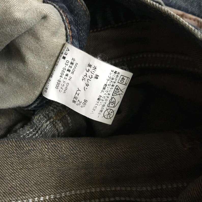 Pu&ograve; includere: Primo piano di un'etichetta di un capo in denim. L'etichetta, stampata in giapponese e inglese, indica la composizione del tessuto: 98% cotone, 2% poliuretano. Sono visibili anche le istruzioni di lavaggio e "MADE IN CHINA".