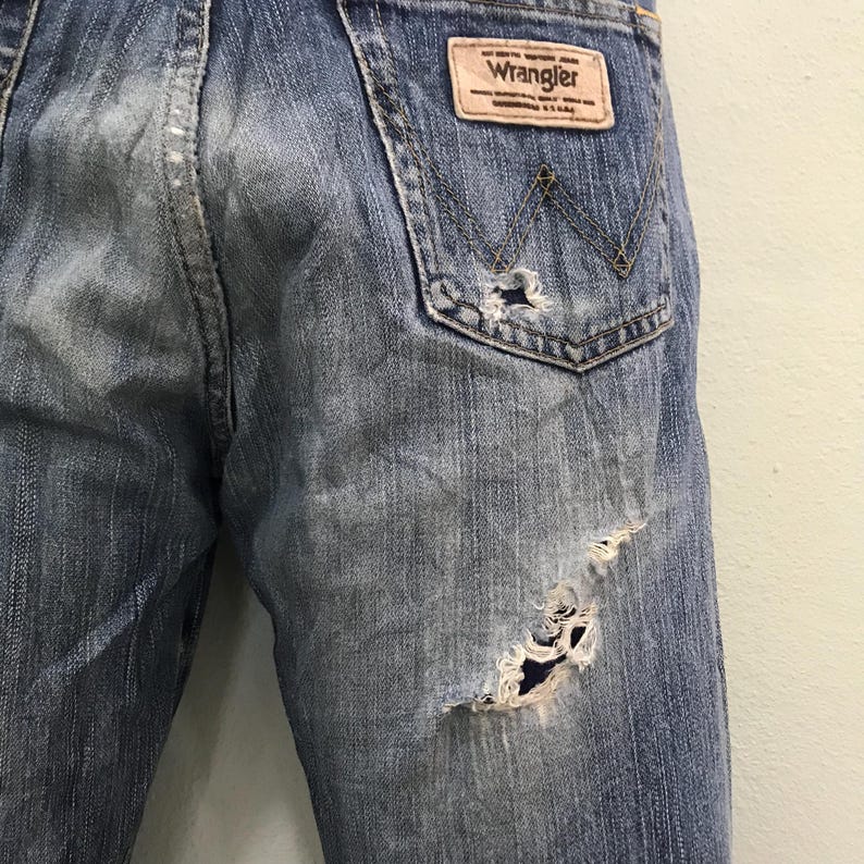 Op de afbeelding: Close-up van een distressed blauwe denim jeans. De jeans heeft een versleten look met scheuren en vervaging. Een bruine leren patch met het "Wrangler"-logo is zichtbaar op de achterzak.