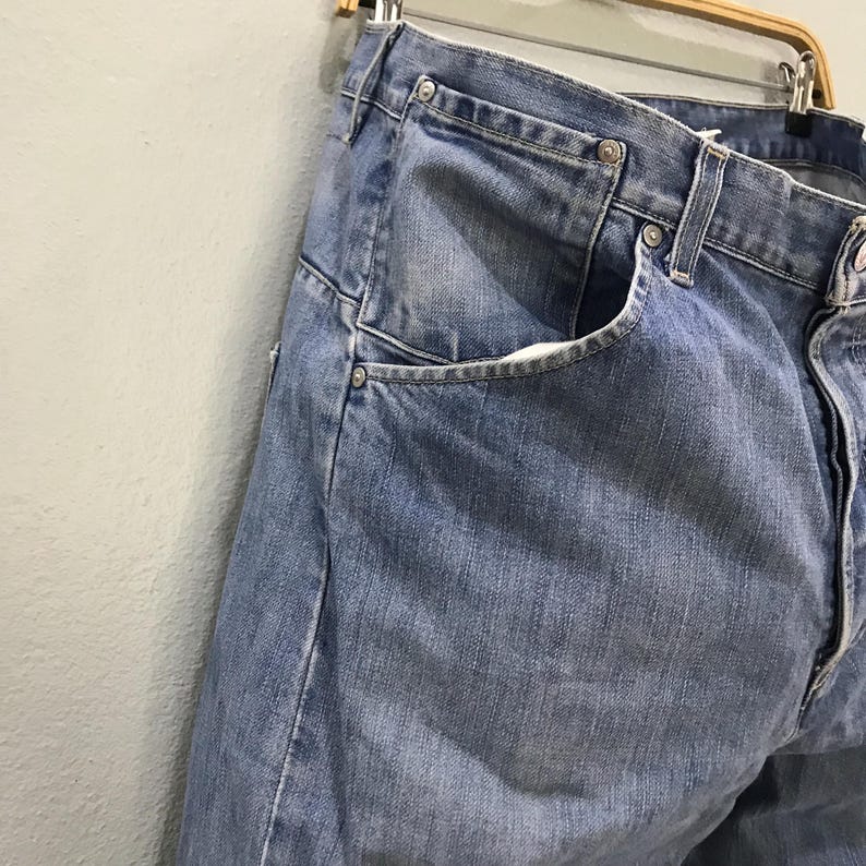 May include: A pair of blue denim jeans featuring a classic five-pocket design with metal rivets at the corners of the front pockets, a button fly closure, and a waistband with belt loops, displayed hanging on a wooden hanger against a wall.