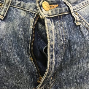 Op de afbeelding: Close-up van een blauwe spijkerbroek met een gedeeltelijk open rits. De jeans heeft een messingkleurige knoop met de merknaam "Wrangler" en een messing rits. De denim heeft een vervaagde, versleten uitstraling.