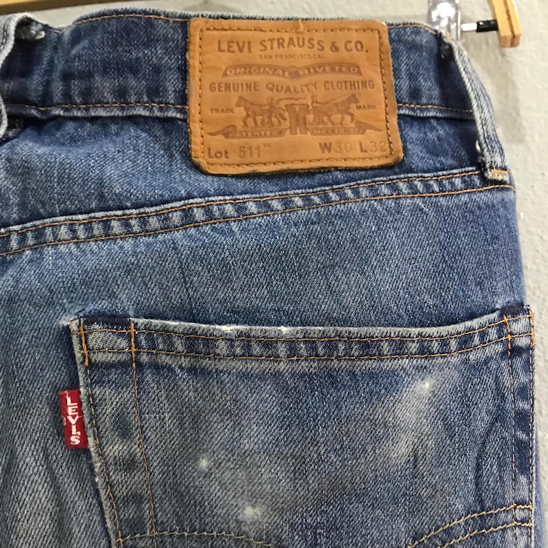 May include: Close-up of blue denim jeans with a brown leather patch featuring the Levi Strauss & Co. logo. The jeans have a back pocket with a red Levi's tag and are labeled Lot 511, W30 L32.