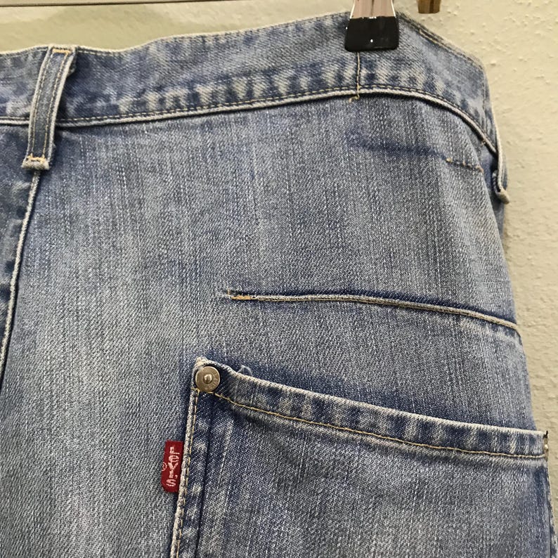 May include: Light blue denim jeans featuring a classic five-pocket construction with a high waistband, belt loops, and a metal rivet at the corner of the pocket. A small red fabric tag with the white text Levi's is attached to the pocket seam.
