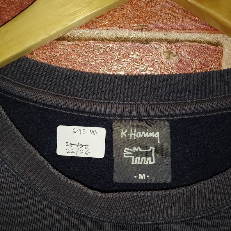 May include: A black sweatshirt with a white tag that says "693 W" and "93/26 22/26". There is also a black tag with the text "K. Haring" and a stylized drawing of a dog. The tag also says "M".