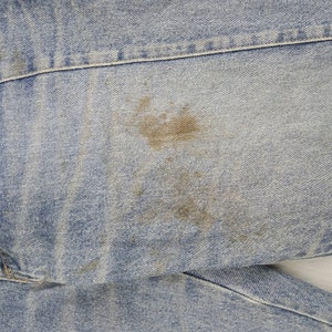 Może przedstawiać: Zbliżenie jasnoniebieskiego materiału dżinsowego z brązową plamą. Dżins ma widoczne szwy i teksturowaną powierzchnię. Plama ma nieregularny kształt i wydaje się być przebarwieniem.