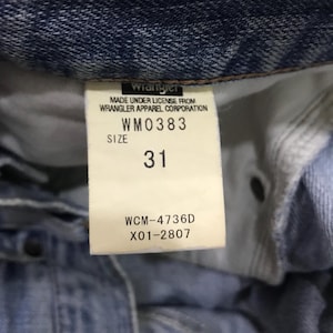 May include: Close-up of a denim garment label. The label reads "Wrangler" and includes the text "MADE UNDER LICENSE FROM WRANGLER APPAREL CORPORATION." The size is indicated as "31" with the style number "WM0383" and other codes.