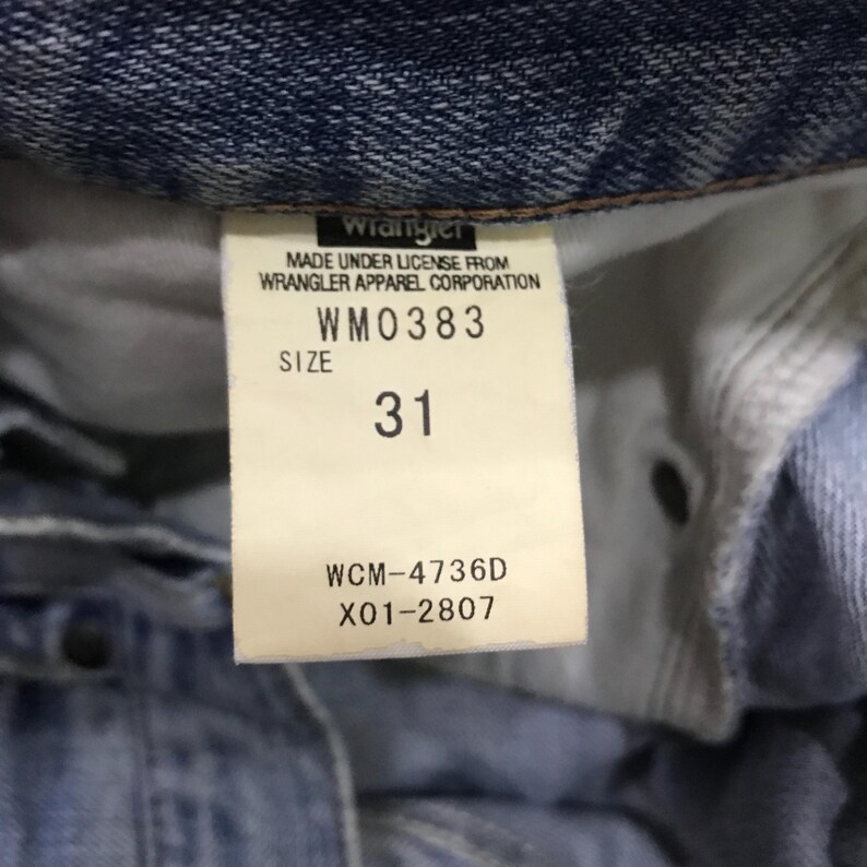 May include: Close-up of a denim garment label. The label reads "Wrangler" and includes the text "MADE UNDER LICENSE FROM WRANGLER APPAREL CORPORATION." The size is indicated as "31" with the style number "WM0383" and other codes.