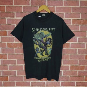 May include: Black t-shirt with a graphic featuring the text "SHADOWFIST" above a colorful illustration of a monster and figures in combat. The text "EQUAL OPPORTUNITY BUTT KICKING" is below the illustration.