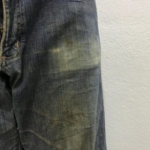 以下が含まれることがあります： 色あせた部分と微妙な変色が見られる、青色のデニムジーンズのクローズアップ。ジーンズは、使用感のある、テクスチャのある外観を示しています。生地は、目に見えるステッチと伝統的なデザインを備えた、クラシックなデニムの外観をしています。