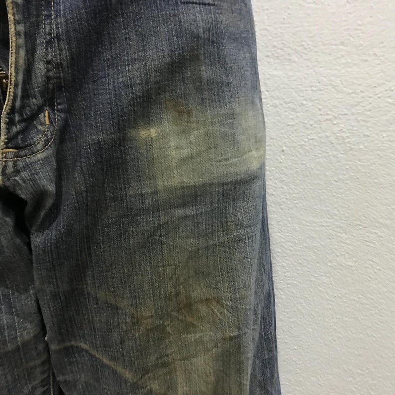 以下が含まれることがあります： 色あせた部分と微妙な変色が見られる、青色のデニムジーンズのクローズアップ。ジーンズは、使用感のある、テクスチャのある外観を示しています。生地は、目に見えるステッチと伝統的なデザインを備えた、クラシックなデニムの外観をしています。