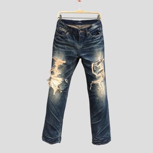 Può includere: Jeans in denim blu slavato con dettagli strappati. I jeans sono appesi a una gruccia di legno su uno sfondo grigio chiaro. I jeans hanno un classico design a cinque tasche e una chiusura con bottone e cerniera.