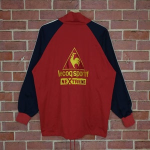 Puede incluir: Chaqueta de ch&aacute;ndal roja, negra y blanca con un logotipo de gallo amarillo y el texto "le coq sportif NEXTREME" en la espalda.