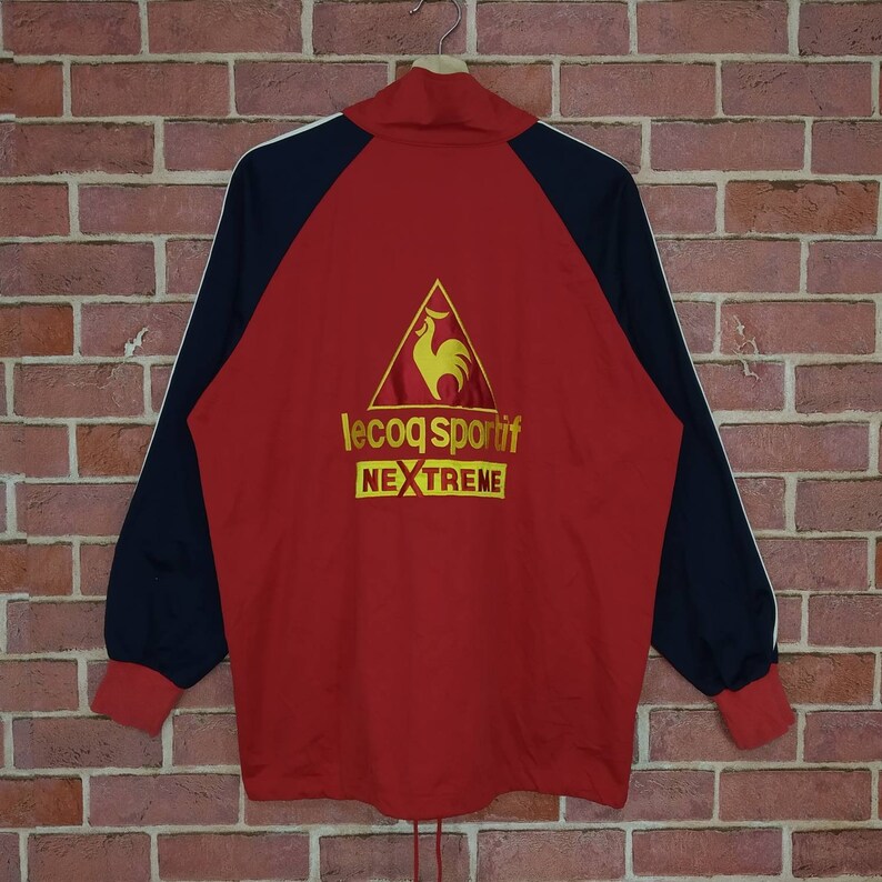 Puede incluir: Chaqueta de ch&aacute;ndal roja, negra y blanca con un logotipo de gallo amarillo y el texto "le coq sportif NEXTREME" en la espalda.