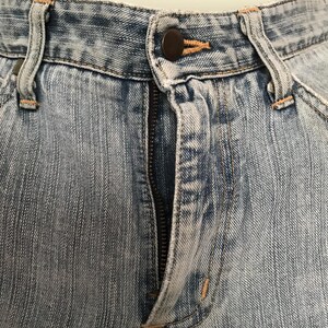 K&ouml;nnte beinhalten: Nahaufnahme einer hellblauen Jeans. Das Bild zeigt den Knopf, den Rei&szlig;verschluss und die G&uuml;rtelschlaufen. Der Denim hat einen verblichenen, abgenutzten Look. Die N&auml;hte sind in einer kontrastierenden orangefarbenen Farbe sichtbar.
