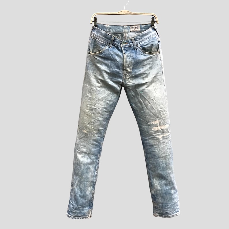 May include: Light blue denim jeans with a faded, distressed appearance. The jeans are hanging on a wooden hanger. The jeans have a classic five-pocket design and a button and zipper closure.