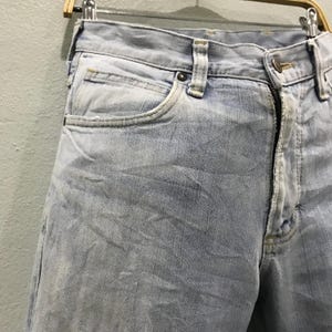May include: Pale blue denim jeans with a faded appearance. The jeans have a traditional five-pocket layout, a button and zip fastening, and belt loops. The fabric shows signs of wear, contributing to a vintage look.
