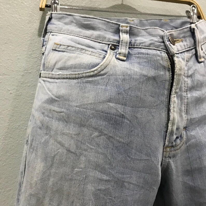May include: Pale blue denim jeans with a faded appearance. The jeans have a traditional five-pocket layout, a button and zip fastening, and belt loops. The fabric shows signs of wear, contributing to a vintage look.