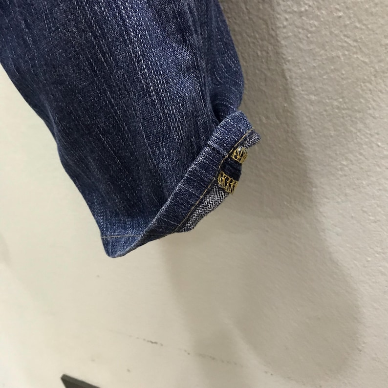 Op de afbeelding: Close-up van een deel van een blauwe spijkerbroek. De manchet is omgeslagen en toont een gedetailleerde naad en decoratieve goudkleurige metalen knopen. De denim heeft een klassieke, versleten uitstraling.
