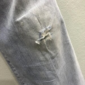 May include: Close-up of light blue denim jeans with a distressed, ripped knee. The fabric shows fading and wear, with frayed edges around the tear. The jeans appear to be pre-worn or intentionally distressed for a vintage look.