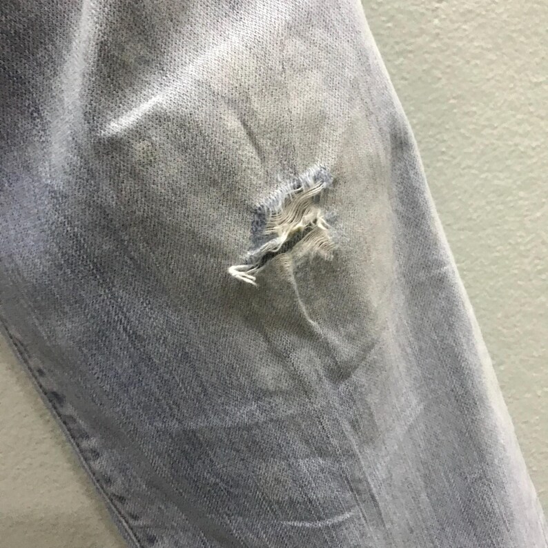 May include: Close-up of light blue denim jeans with a distressed, ripped knee. The fabric shows fading and wear, with frayed edges around the tear. The jeans appear to be pre-worn or intentionally distressed for a vintage look.